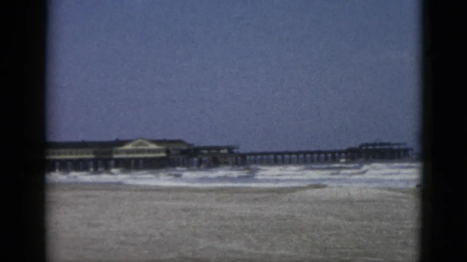 8mm home-movie frame, circa 1952, Quebec beach with wooden pier extending across mid-frame and pavilion building at centre.