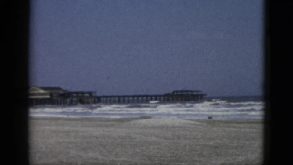 8mm home-movie frame, circa 1952, Quebec beach with long wooden pier, pavilion structure, and empty sandy foreground in the closing seconds of the clip.