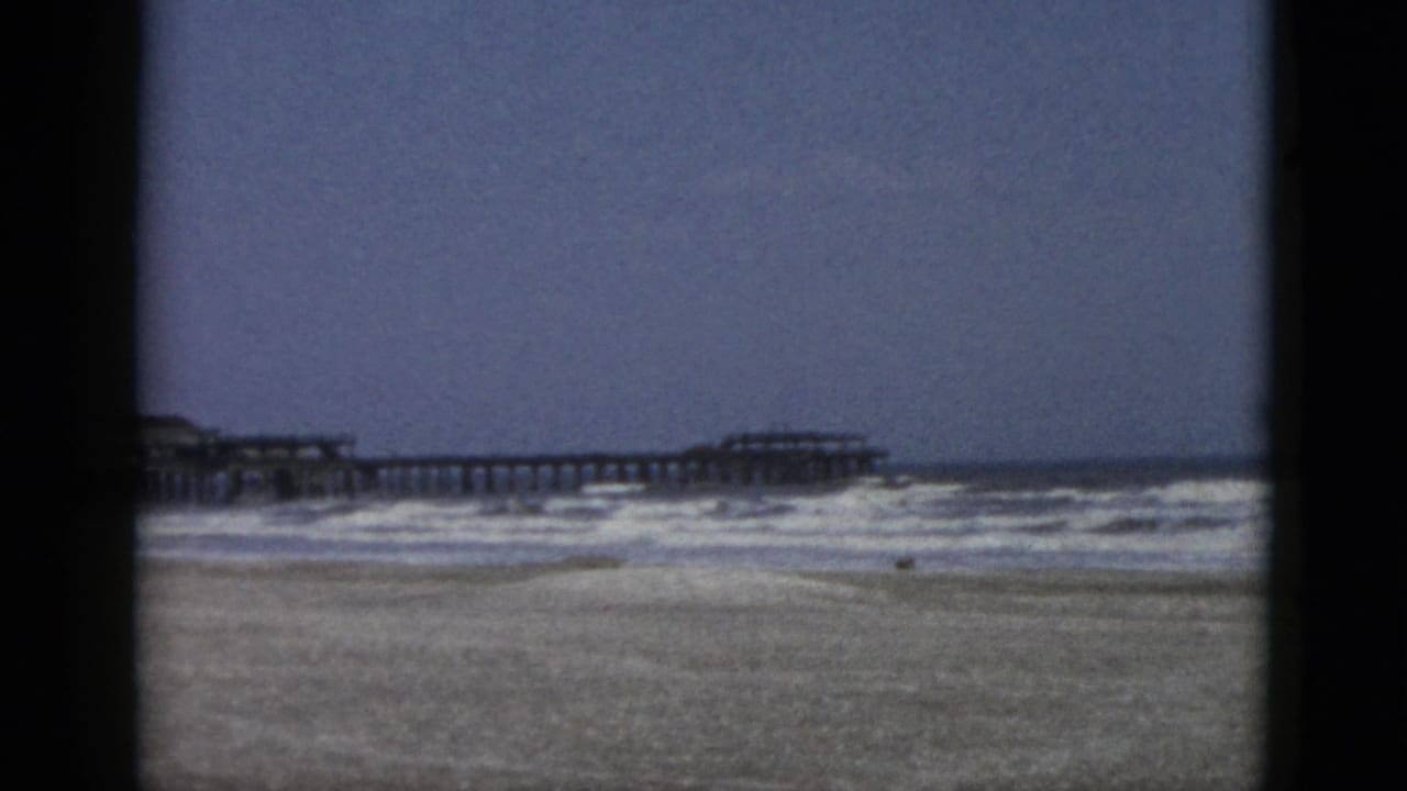 Sandy beach with breaking waves and a long wooden pier extending into the sea under a grey-blue sky.