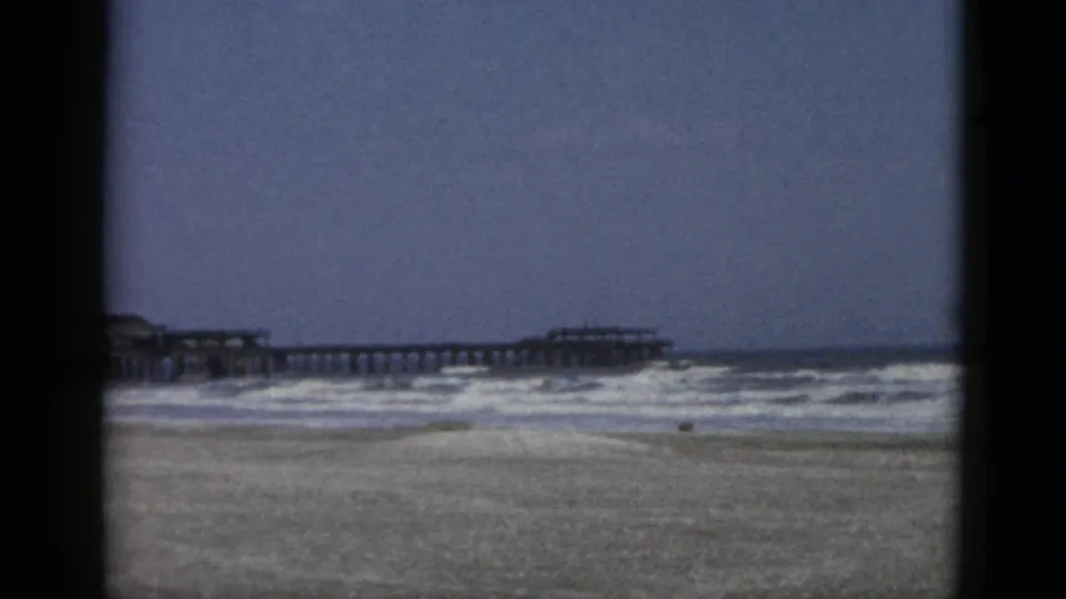 8mm home-movie frame circa 1952 showing a sandy shoreline with white-capped surf and a pier structure extending seaward under an overcast sky.