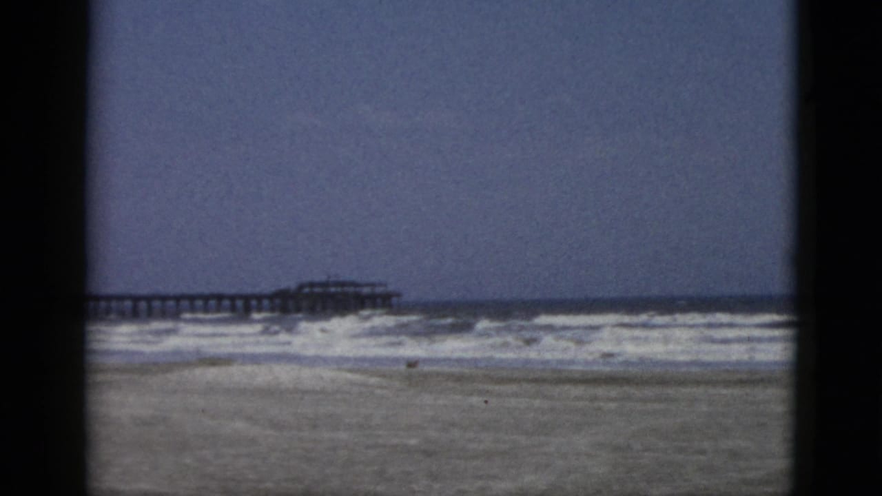 Second 8mm frame in a circa-1952 beach sequence showing surf, wet sand, and a pier extending into the sea under a hazy sky.