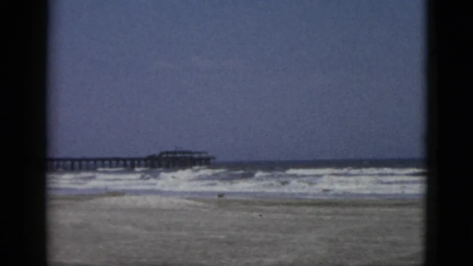 Second 8mm frame in a circa-1952 beach sequence showing surf, wet sand, and a pier extending into the sea under a hazy sky.