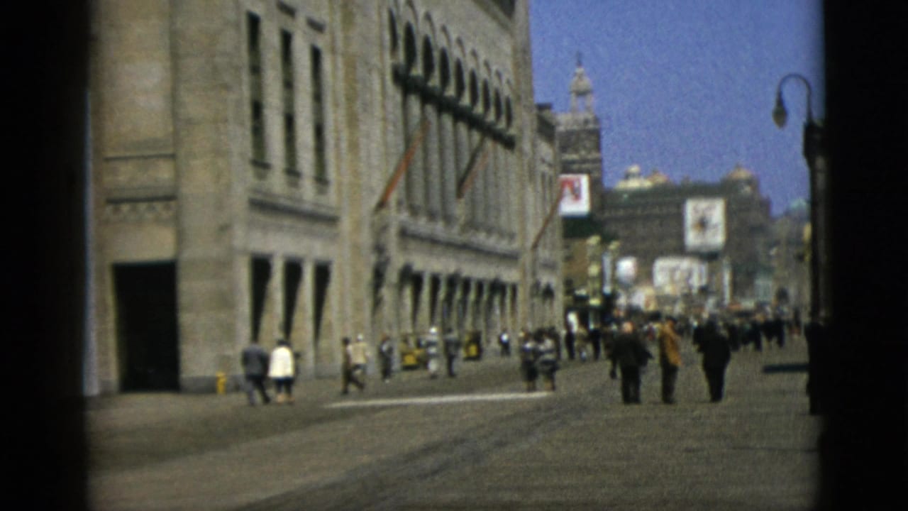 Pedestrians in mid-century attire walk a wide urban boulevard flanked by grand stone neoclassical buildings, advertising billboards visible in the distance.