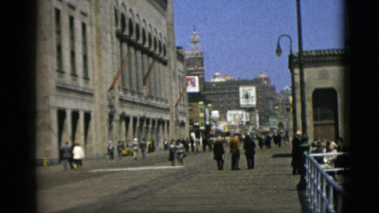 Quebec, Canada, 1952 — 8mm frame showing pedestrians on a wide boulevard with stone facades on both sides, a metal railing, globe street lamp, distant tower, and advertising billboards.