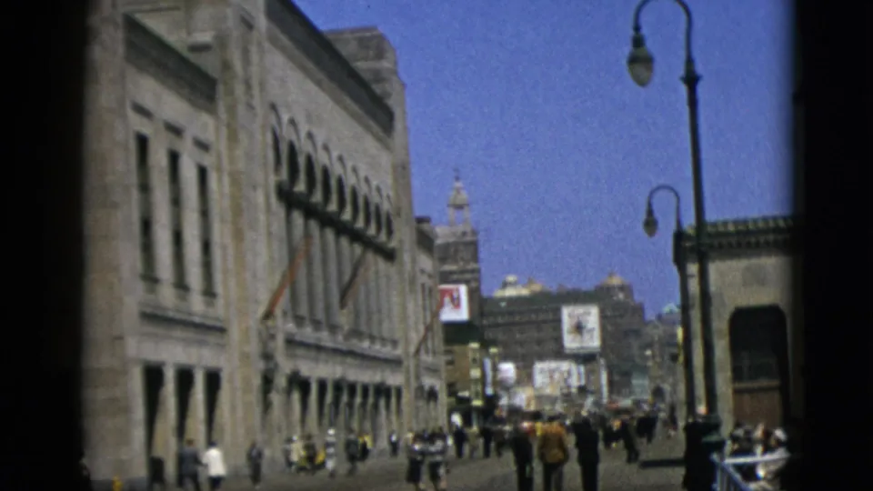 Quebec, Canada, 1952 — 8mm frame emphasizing a grand stone building facade with arched windows on the right, a globe street lamp in the foreground, and pedestrians receding down the boulevard.