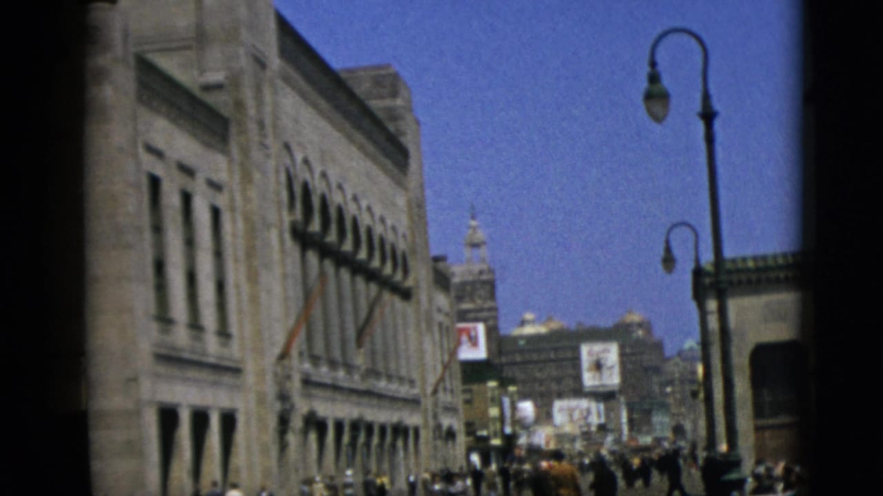 Street-level view of a large stone building with arched windows; a lamppost stands center-frame with a crowd visible at street level and illuminated signs in the distance.