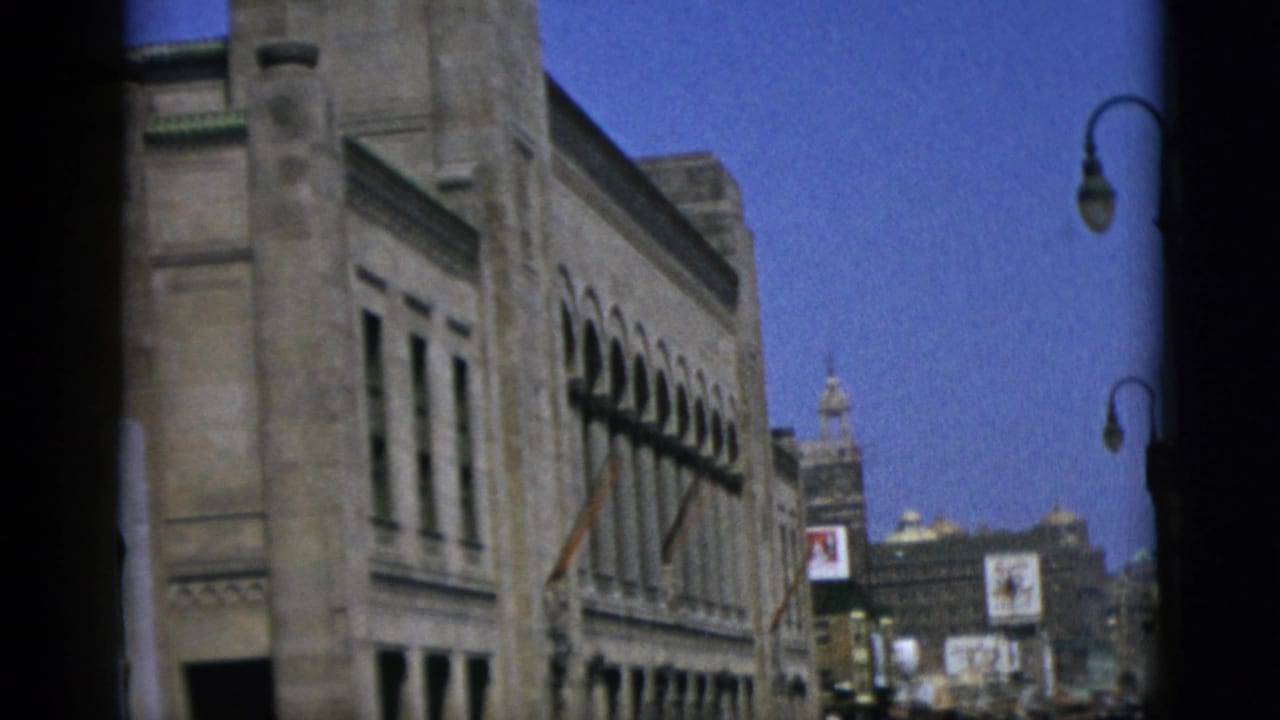 Classical stone building facade with arched windows; lamppost at right edge; illuminated signs and a tower visible in the distance against a blue sky.