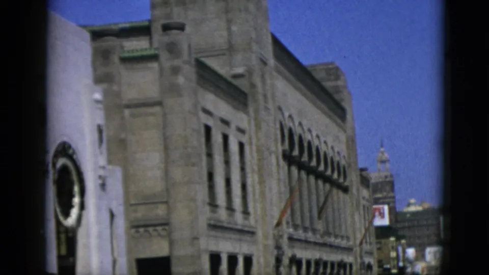 1950s Canadian city street: close view of classical masonry building with arched windows, a circular decorative roundel at ground level, and a tower in the distance under blue sky.