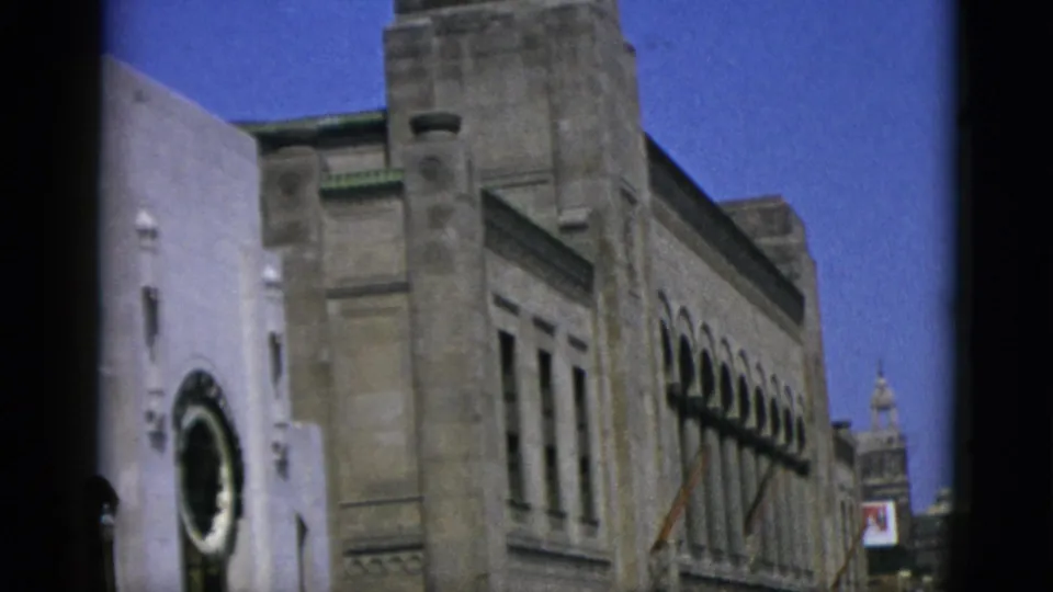 1950s Quebec street scene: classical stone building facade with a circular roundel, arched windows, and a tower in the mid-distance against a saturated blue sky.