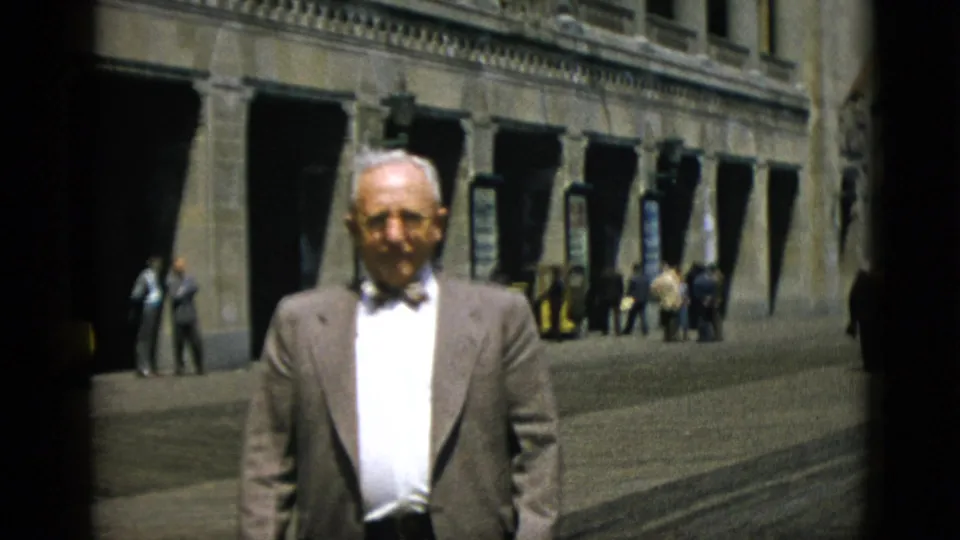 Older man wearing a suit jacket and bow tie poses in front of a large stone public building, Quebec, Canada, 1952, with several pedestrians visible behind.