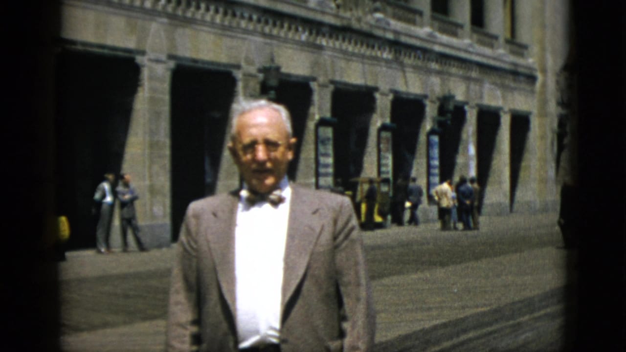 Elderly man in light suit and bow tie stands before a large neoclassical stone building with arched arcade; pedestrians and a yellow vehicle visible behind him.