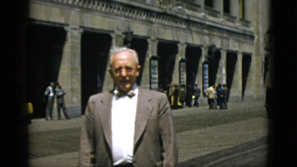 Elderly white-haired man in bow tie poses before a neoclassical stone arcade building in Quebec, Canada, circa 1952; mid-century pedestrians and a yellow vehicle in the background.