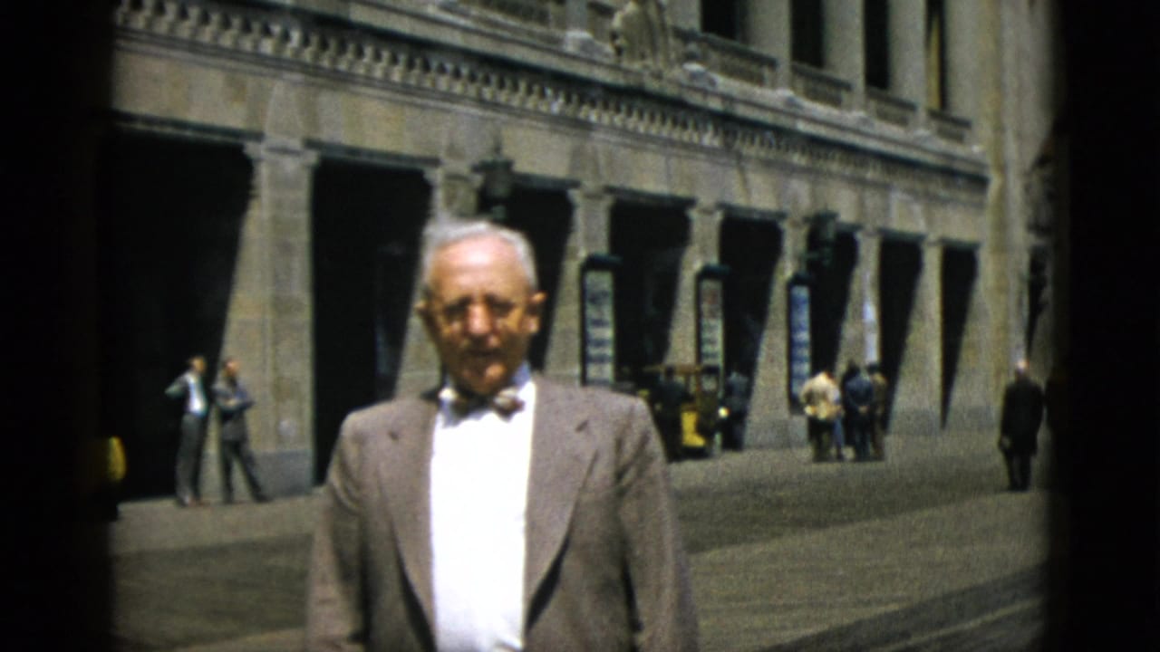 Closer framing of elderly man in bow tie before the stone building arcade in Quebec, Canada, circa 1952; background figures and yellow vehicle still present.