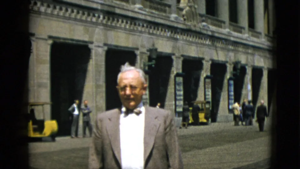 Elderly man in bow tie and suit jacket before Quebec stone building arcade, circa 1952; a yellow vehicle is more prominently visible in the lower background.