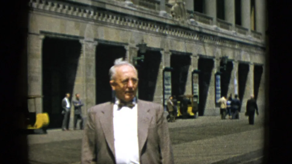 Final frame: elderly man in bow tie before neoclassical stone arcade in Quebec, Canada, circa 1952, with mid-century pedestrians and yellow vehicle behind him.