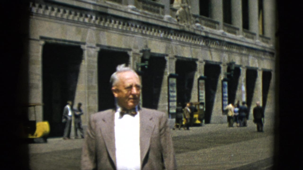 Man in suit and bow tie walks toward camera on a downtown Quebec street, circa 1952, with a large stone classical building and background pedestrians visible.