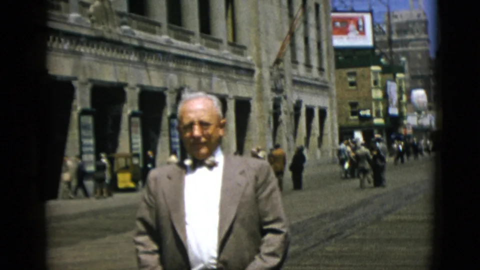 Closer shot of older suited man walking toward camera on a Quebec street, circa 1952, with a billboard advertising sign, yellow vehicle, and pedestrian crowds behind.