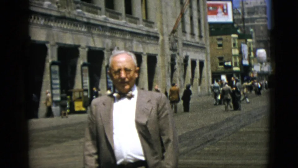 Final frame of older suited man on a Quebec downtown street, circa 1952, facing camera with busy sidewalk crowds and period advertising signs behind him.