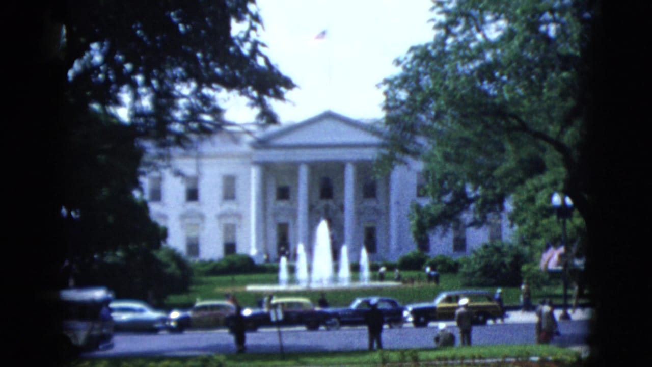 White House North Portico in Washington DC, circa 1952, seen through foreground tree branches, with active fountains and period automobiles at the fence line.