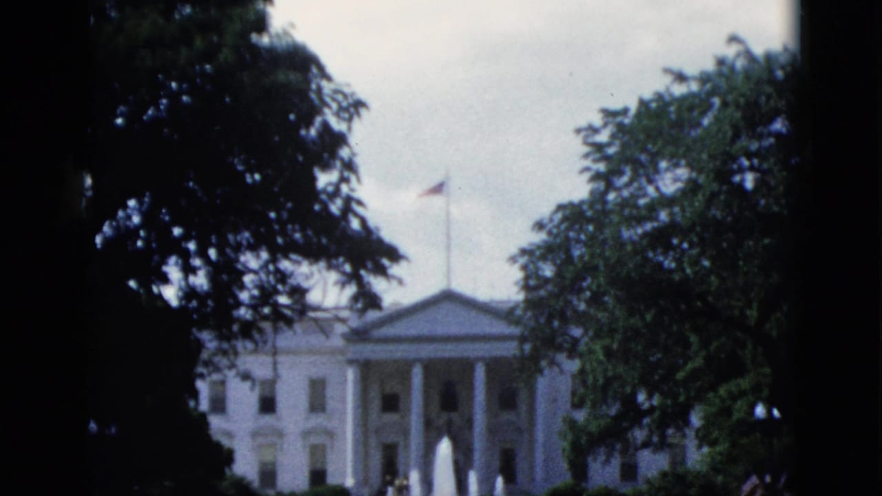 White House north facade framed by dark tree silhouettes, with active fountains and American flag visible, viewed from street level.