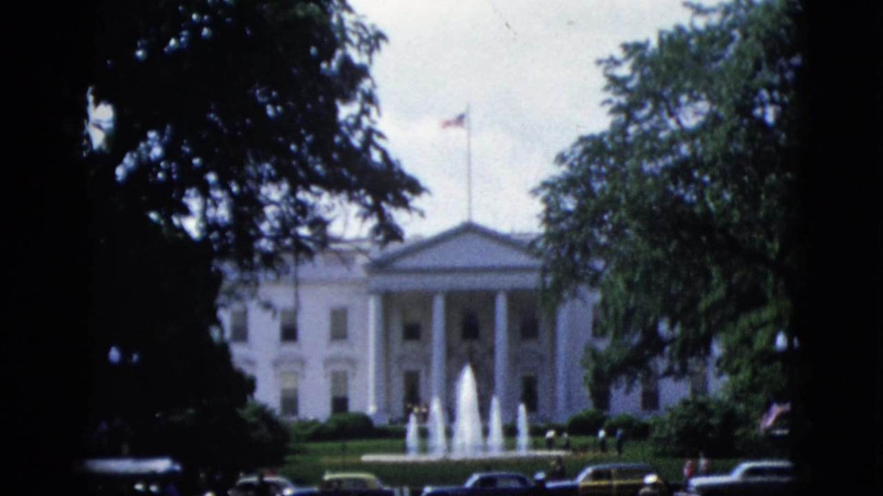 White House north facade with trees left and right, active fountains in front, American flag flying, dark cars partially visible along street at bottom.