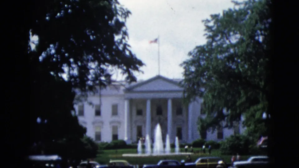 White House north portico viewed through trees circa 1952, active north lawn fountains, American flag, and dark-bodied automobiles parked along the street, 8mm color film.