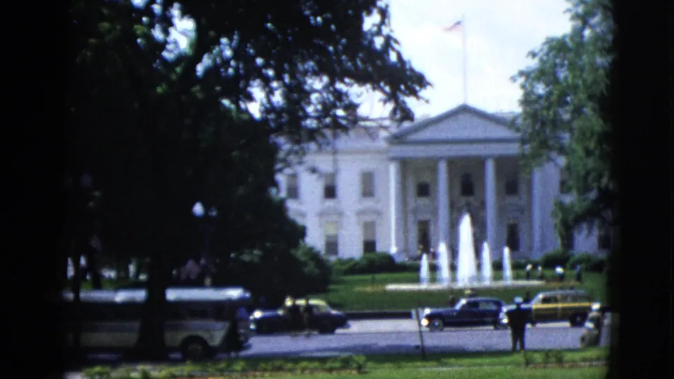 White House north portico circa 1952 viewed through trees, active fountains, dark early-1950s sedans at street level, and two standing figures at lower right, 8mm color film.