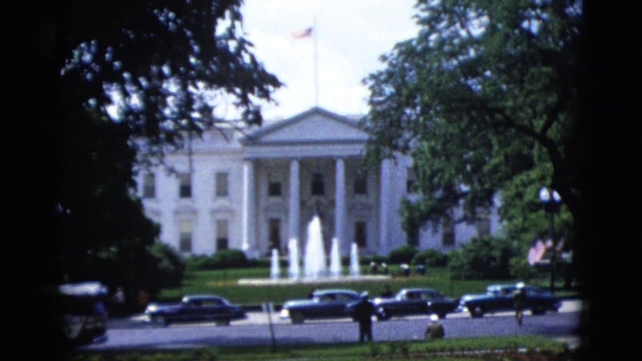 White House North Portico with active fountains and mature trees; a row of classic dark sedans lines the street and an American flag flies on the roofline.