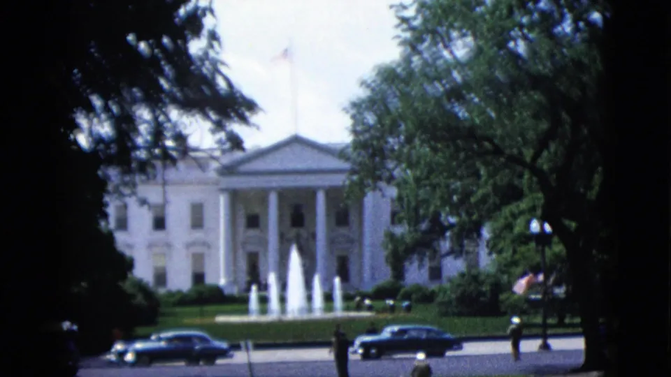 8mm color frame from 1952, Washington DC — White House North Portico with columns and pediment, North Lawn fountain, prominent mature trees, American flag on a pole, and 1950s automobiles on the street.