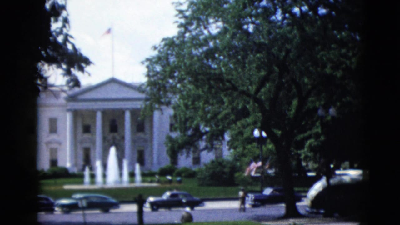 White House North Portico seen through mature trees, with dark sedans and pedestrian figures on the street below, 8mm film.