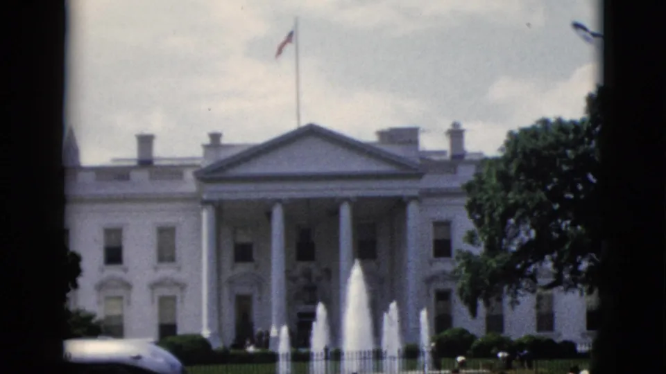White House North Portico, Washington DC, circa 1952 — American flag on roofline, multi-jet fountain, partly cloudy sky, dark car at lower left.