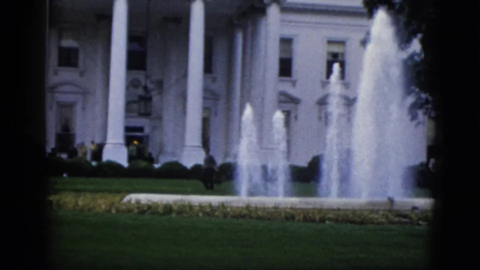 8mm film frame of White House north facade, Washington DC, circa 1952: three fountain jets, columns, flag at roof flagpole, lawn, and small distant figures under overcast sky.