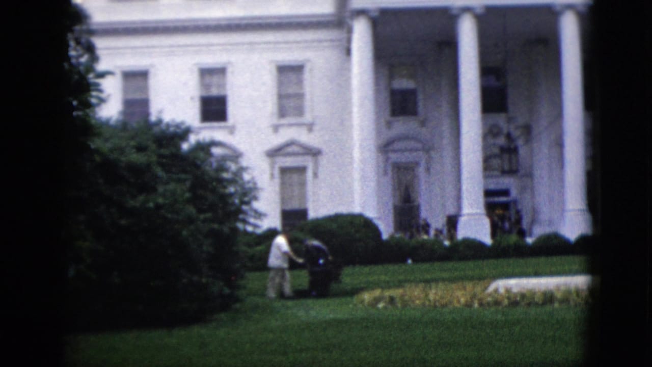 8mm color footage circa 1952 of the White House north portico with a groundskeeper operating a push lawnmower on the lawn; trimmed hedges and dark foliage frame the right edge.