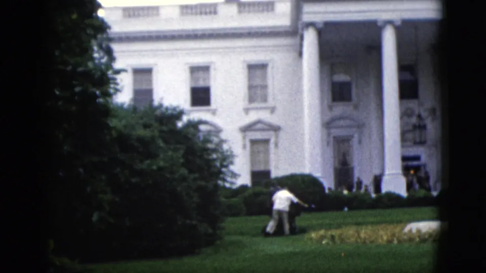 8mm color footage circa 1952 showing the White House north portico columns and a groundskeeper working near the hedge border, with the entrance lantern visible and dark foliage at frame left.