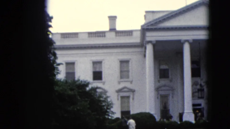 8mm color footage circa 1952 showing the full width of the White House north facade against a pale overcast sky, with a figure near the entrance and foliage at the right frame edge.