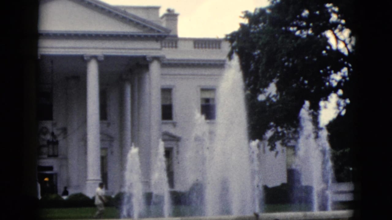 8mm home movie frame, c.1952, White House north facade with active North Lawn fountain, overcast sky, and a lone figure on the lawn.