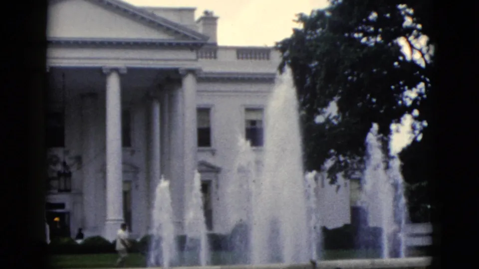 8mm home movie frame, c.1952, White House north facade with active North Lawn fountain, overcast sky, and a lone figure on the lawn.