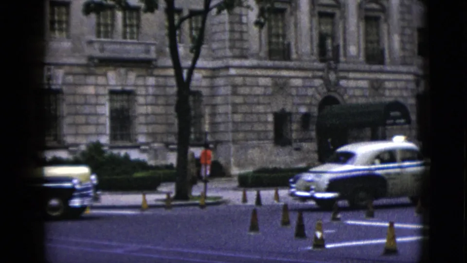 Washington DC street circa 1952: two-tone taxi and dark 1950s sedan pass a neoclassical stone building; orange traffic cones in road; pedestrian figure visible on sidewalk.