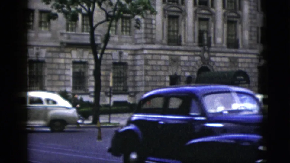 Washington DC street circa 1952: large dark blue 1950s sedan and cream-colored sedan in traffic; neoclassical stone building with arched windows in background.