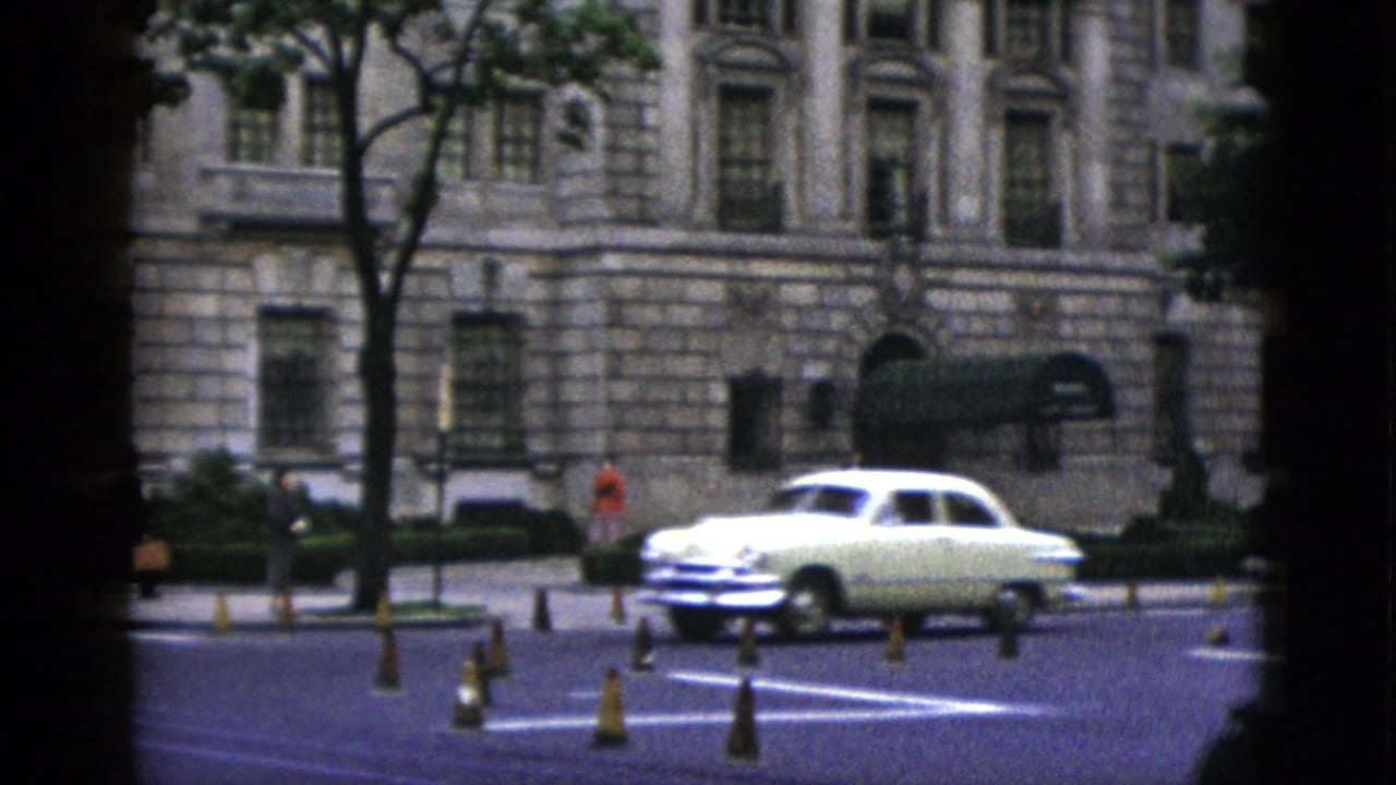 Early-1950s cream-colored American sedan at a Washington DC intersection, neoclassical civic building with green awnings behind, orange traffic delineators in roadway.