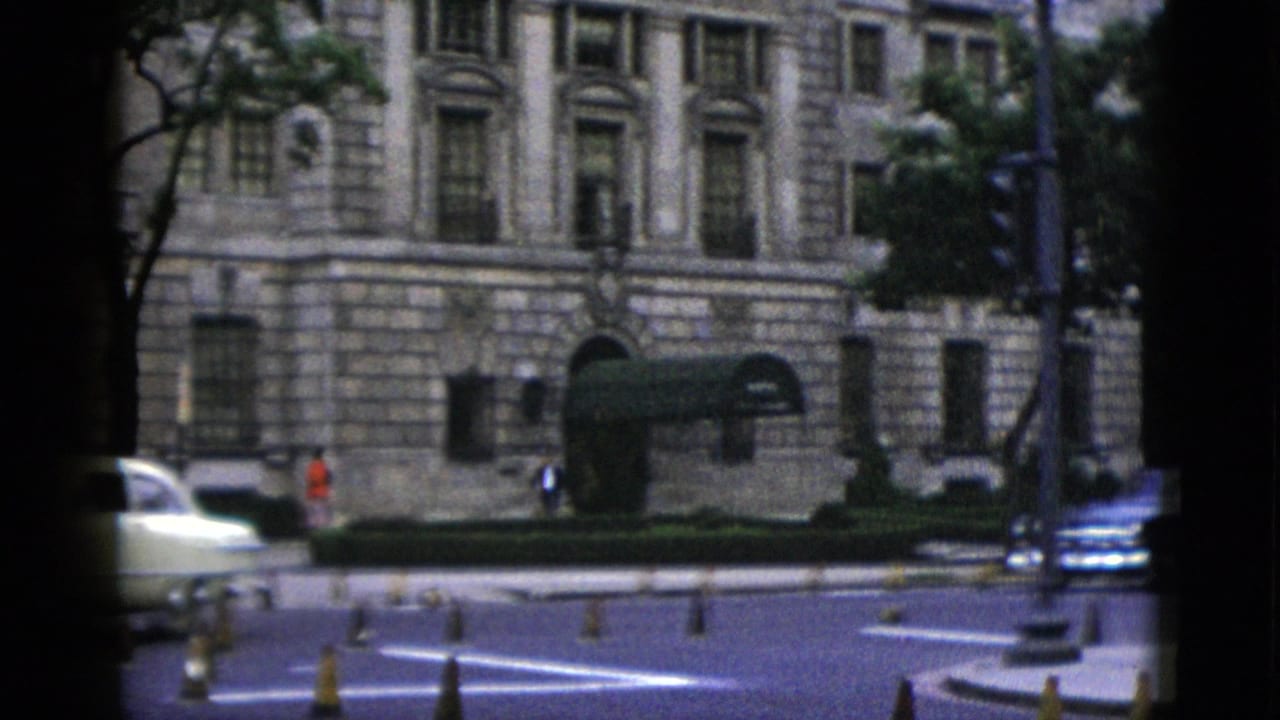 Mid-century American sedan crossing a Washington DC intersection; neoclassical building facade with green awnings and period traffic signal visible.
