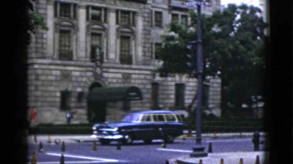 Dark 1950s American station wagon navigating a Washington DC intersection; neoclassical civic building with stone columns and mature street trees in background.