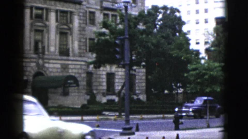 Motion-blurred cream-colored 1950s sedan crossing an intersection near a large neoclassical civic building in Washington DC; dark vehicle at right.