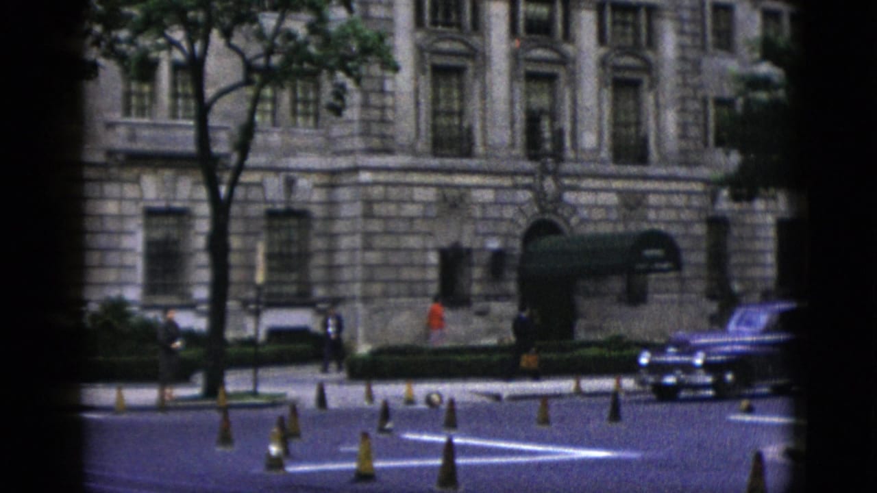 Wide street view of a large neoclassical stone building with dark awnings; early 1950s sedans and orange traffic cones line the roadway in front.