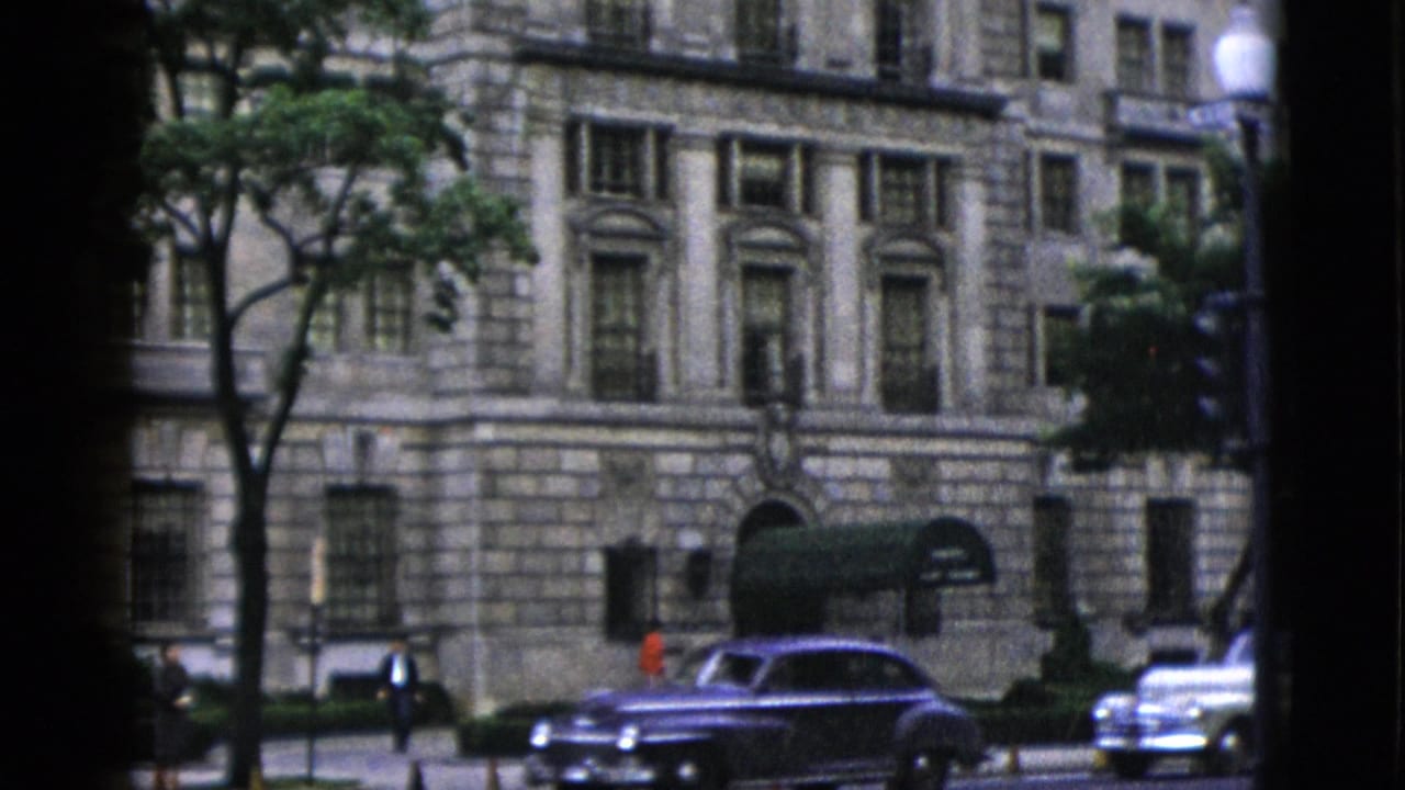 1952 Washington DC — neoclassical stone building with arched doorways and dark awnings; early 1950s American sedans navigate a wide street lined with orange traffic cones, shot on 8mm color film.