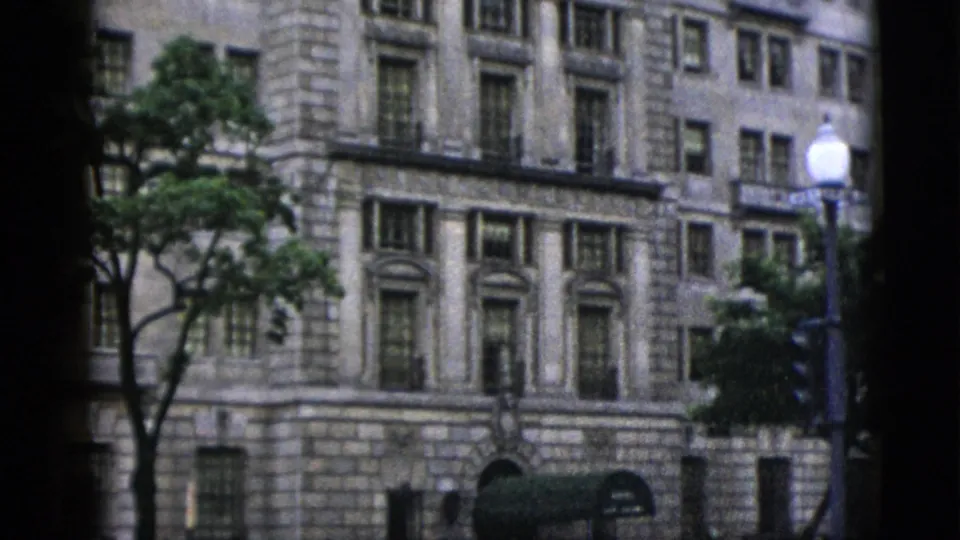 1952 Washington DC — neoclassical stone building with columned facade, white globe street lamp, and traffic signal; summer foliage partially frames the view, shot on 8mm color film.