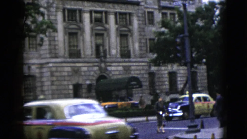1952 Washington DC — a yellow-and-red American sedan occupies the foreground as additional 1950s vehicles and pedestrians pass in front of a neoclassical stone building, shot on 8mm color film.