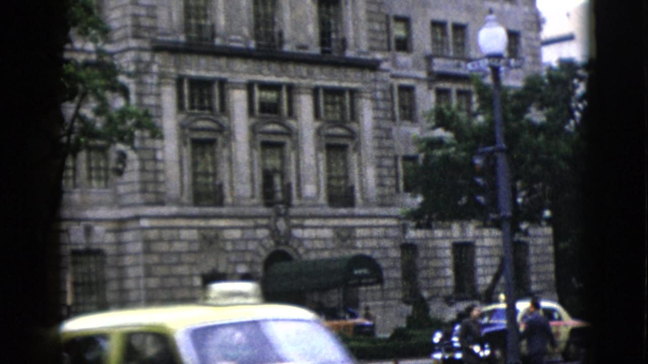 Yellow taxi and dark sedan pass before a multi-story neoclassical stone building in Washington DC, circa 1952, with pedestrians visible at street level.