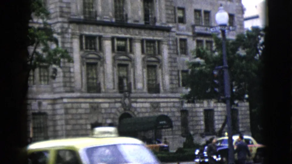 Yellow taxi and dark sedan pass before a multi-story neoclassical stone building in Washington DC, circa 1952, with pedestrians visible at street level.