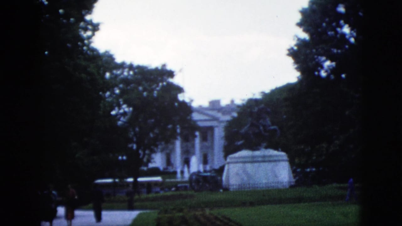 White House facade through trees with equestrian statue pedestal right of center, walking figures on the path in the foreground.